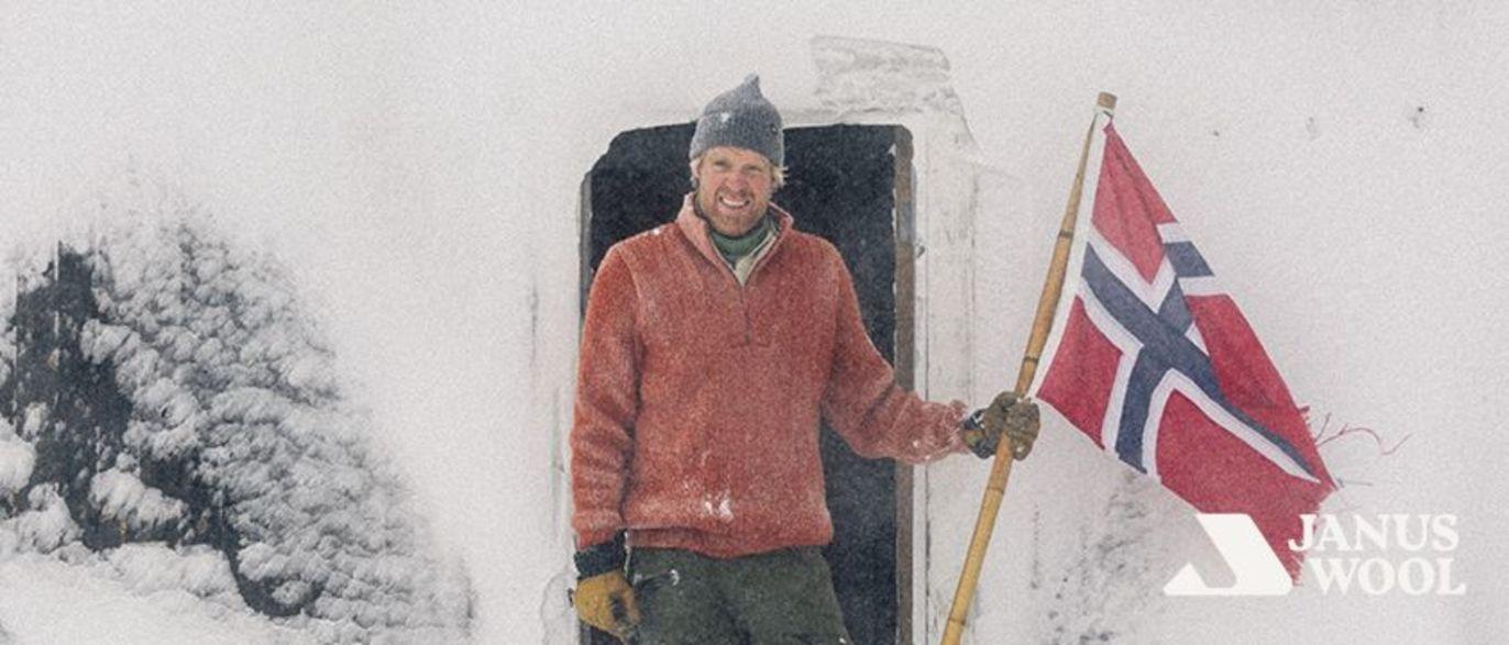 Mann i snøvær som holder det norske flagget utenfor en hytte, iført en rød genser og lue.