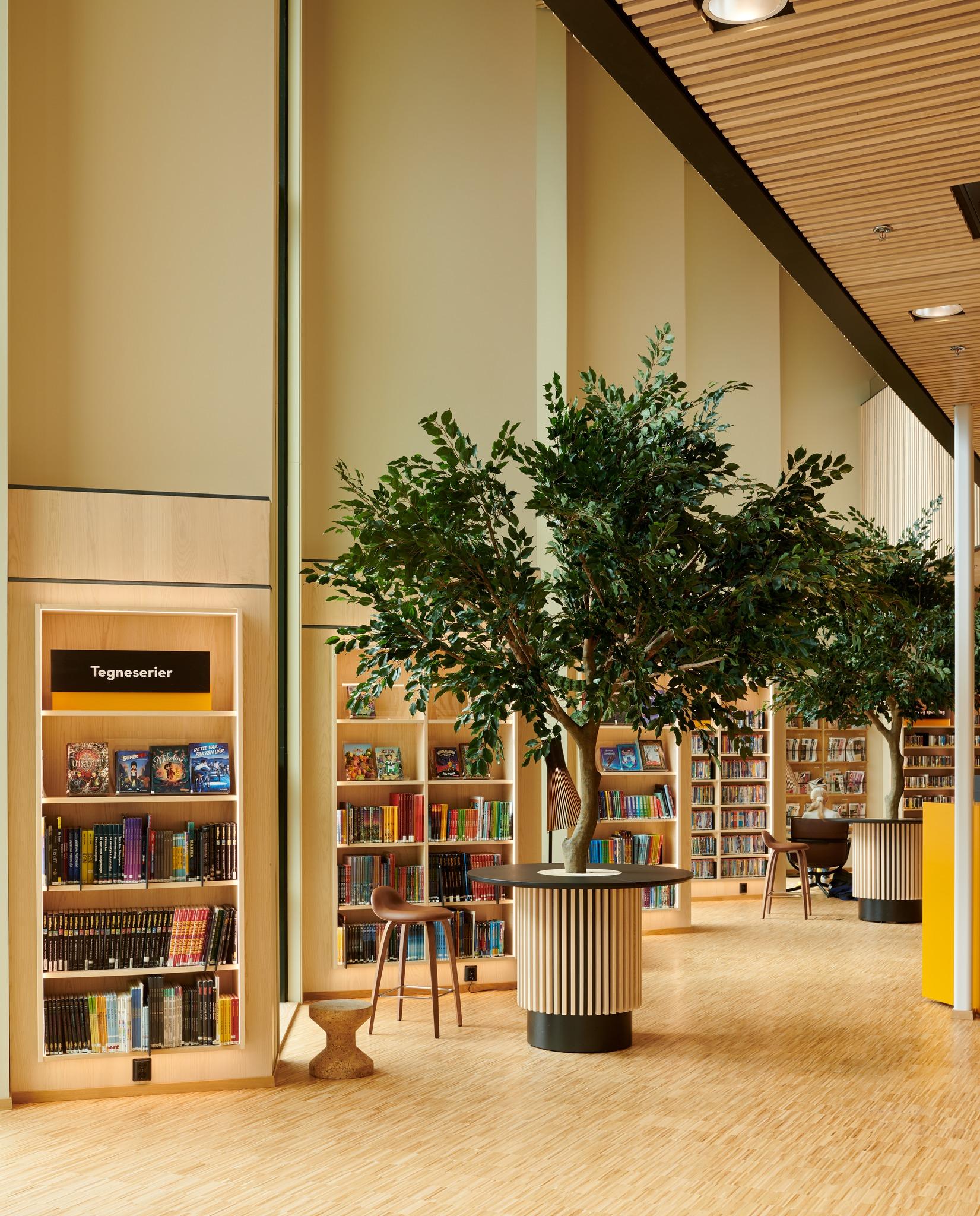 Moderne bibliotekinnredning med bokhyller, grønne trær og lys, som skaper en innbydende atmosfære.