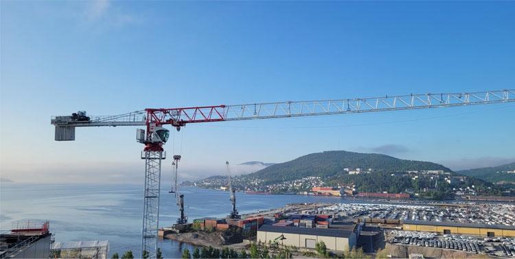 Kran som står over havnen med bygninger og fjell i bakgrunnen, under en klar blå himmel.
