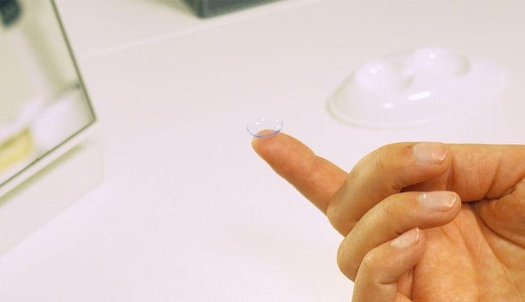 En hånd som holder en kontaktlinse på en finger, med en speil og linsebeholder i bakgrunnen.