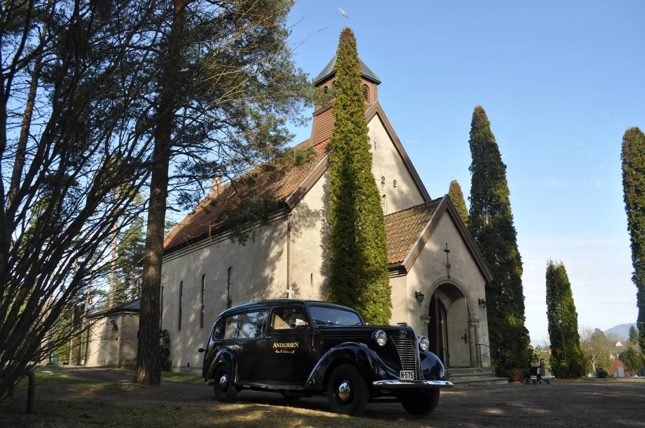 En svart vintage bil parkert foran en kirke omkranset av høye trær og busker.