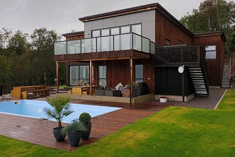 Moderne hus med terrasse, svømmebasseng og hage, omgitt av trær og grønt gress.