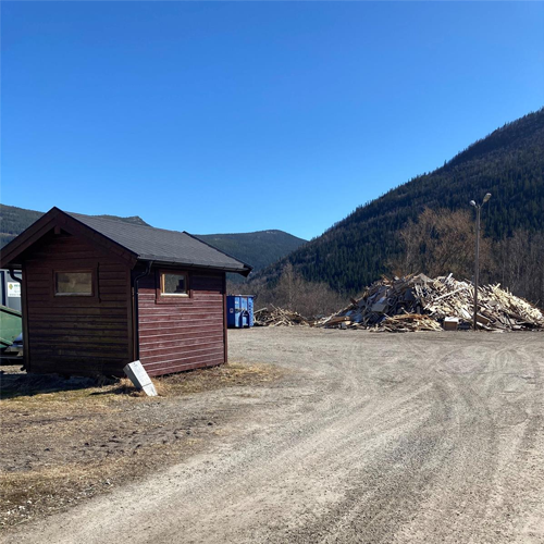 Liten rød hytte ved en grusvei, med fjell og en haug med trevirke i bakgrunnen. Klart blå himmel.