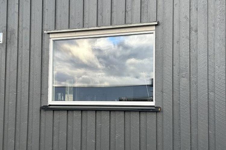 Vindu med refleksjon av skyer på en grå vegg av tre.