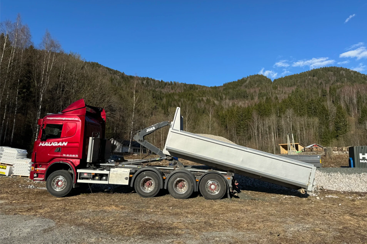 En rød lastebil med tippvogn som tømmer grus på en byggeplass med fjell i bakgrunnen.