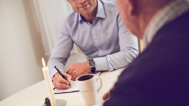 To menn sitter ved et bord, den ene skriver notater mens den andre ser på, med en kopp kaffe og et stearinlys.