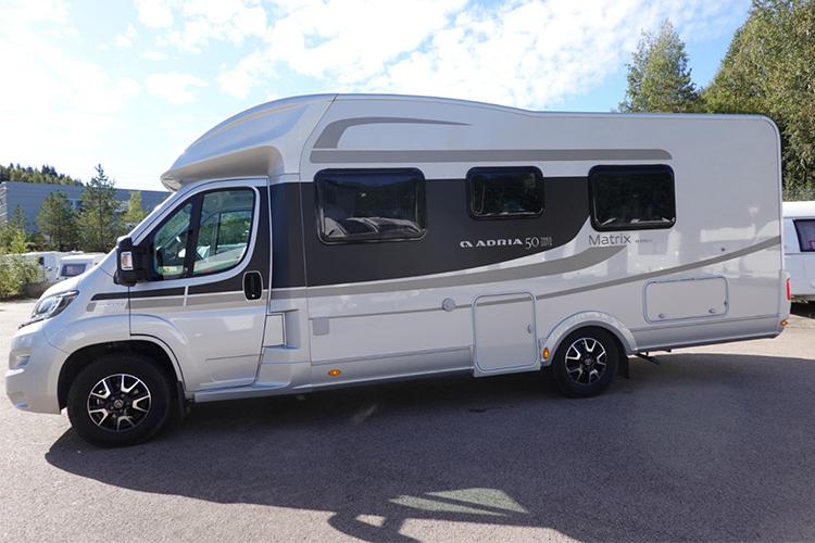 En sølvfarget campingvogn med tre vinduer, parkert på en asfaltvei med trær i bakgrunnen.