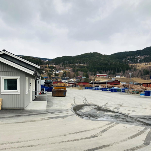 Utsikt over en parkeringsplass med bygninger og fjell i bakgrunnen, delvis overskyet himmel.