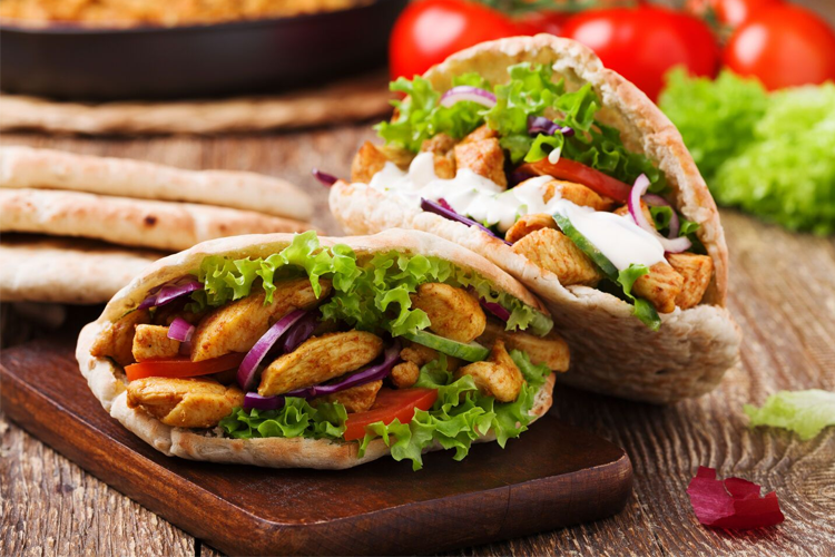 Frisk pita med kylling, salat, tomat og dressing, servert på en treplate med grønnsaker i bakgrunnen.