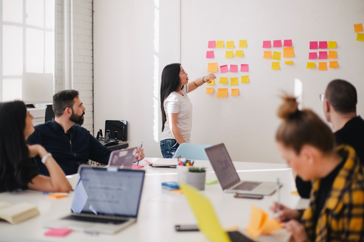 En kvinne skriver på en tavle med fargerike post-it-lapper, mens kolleger ser på i et moderne møterom.