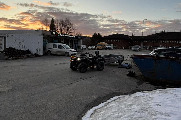 En firehjuling trekker en tilhenger på en parkeringsplass med snø og bygninger i bakgrunnen.