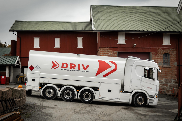 En hvit tankbil med DRIV-logo parkerer ved en rød låve i et gårdsområde.