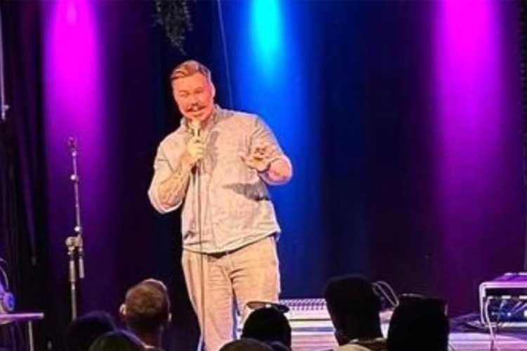 En komiker står på scenen med mikrofon, omgitt av fargerike lys under en stand-up forestilling.