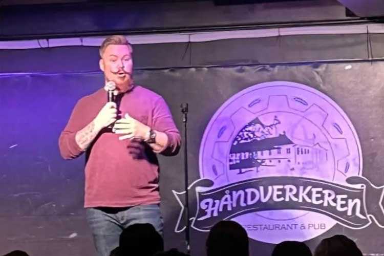 En komiker står på scenen med mikrofon i hånden, foran en logo for Håndverkeren restaurant og pub.