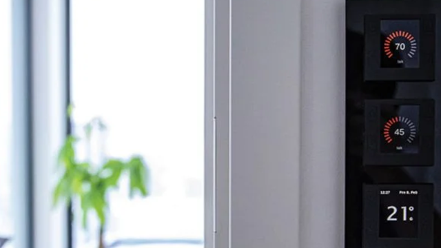 Termostat på veggen viser temperatur og innstillinger i et moderne rom med planter i bakgrunnen.