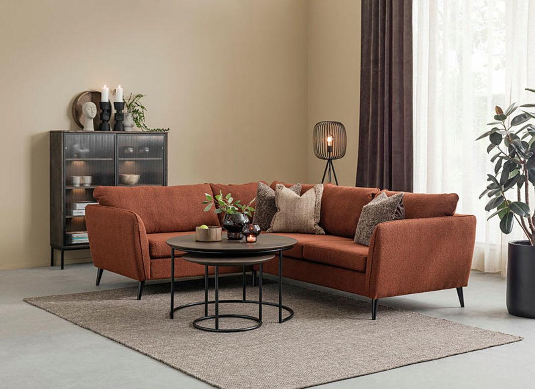 Moderne stue med en rustfarget sofa, et rundt bord, planter og dekorative elementer på en lys vegg.