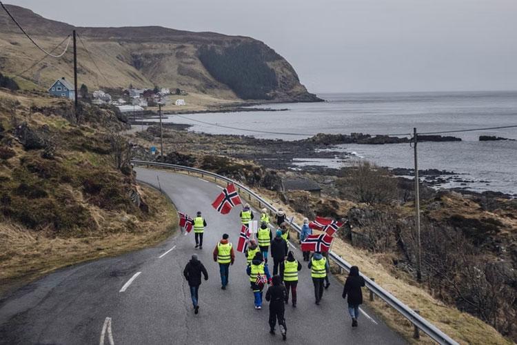 Demonstranter med norske flagg går langs en vei ved kysten, iført gule vester, med fjell i bakgrunnen.