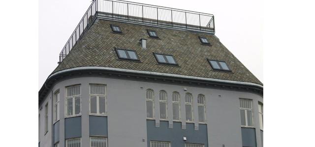 Tak på en grå bygning med skråtak, vinduer og en terrasse på toppen.