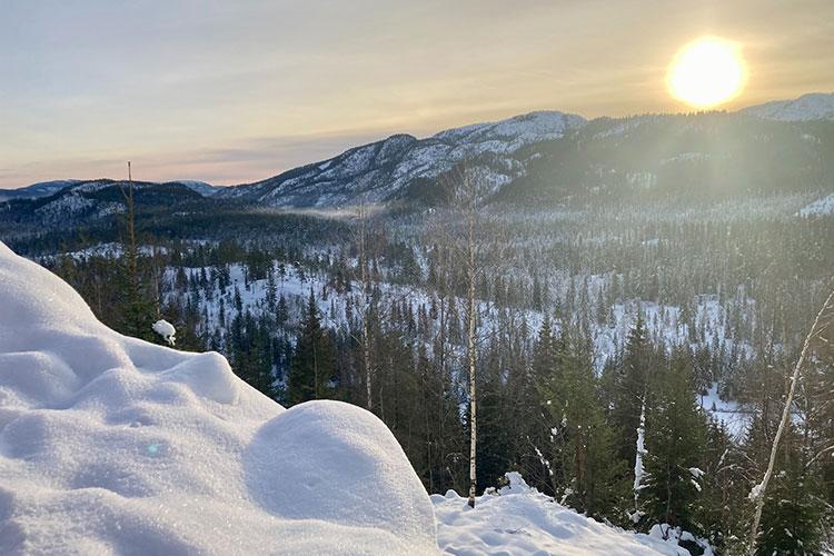 Vinterlandskap med snødekte fjell, trær og en soloppgang som lyser opp himmelen.