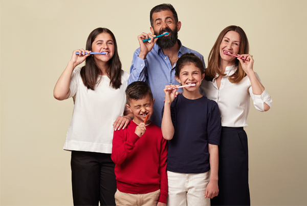 Familie med fem medlemmer som smiler og pusser tennene med tannbørster i hendene.