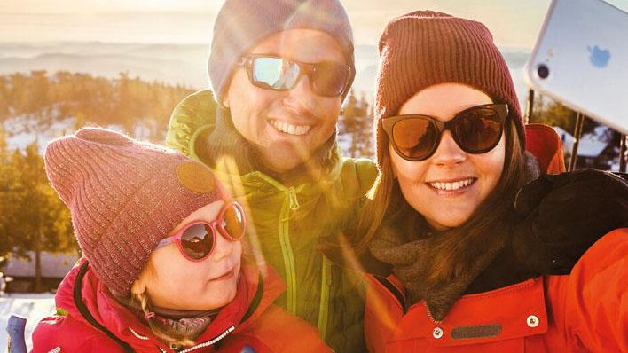 Familie tar selfie i solfylt vinterlandskap, iført luer og solbriller, med snødekte fjell i bakgrunnen.