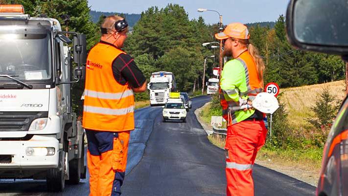 To menn i arbeidsklær står på en vei, mens lastebiler og biler passerer i bakgrunnen.