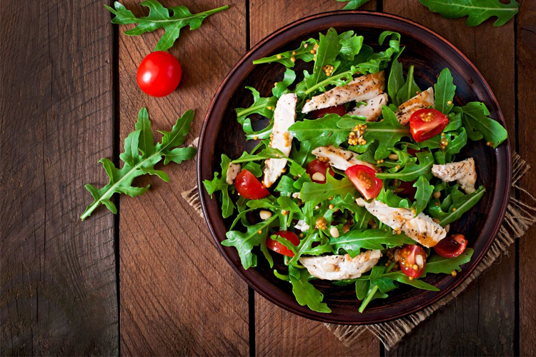 En salat med ruccola, kyllingbiter, cherrytomater og nøtter i en mørk skål på et trebord.
