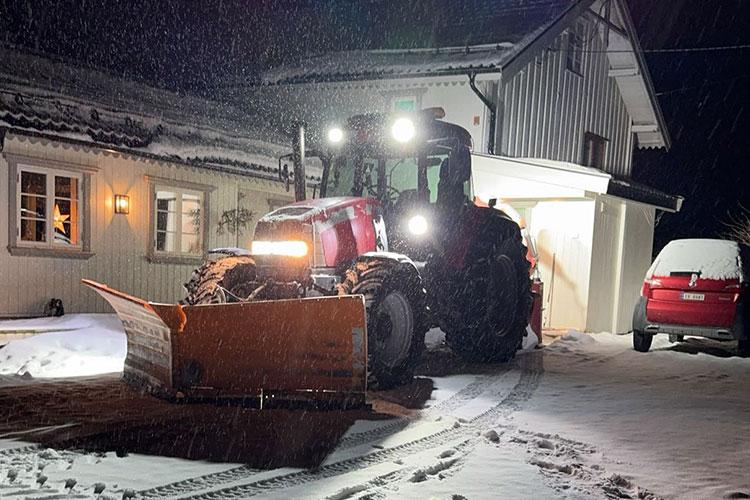 En rød traktor med snøplog står foran et hus i snøvær om natten. Belysningen fra traktoren lyser opp området.