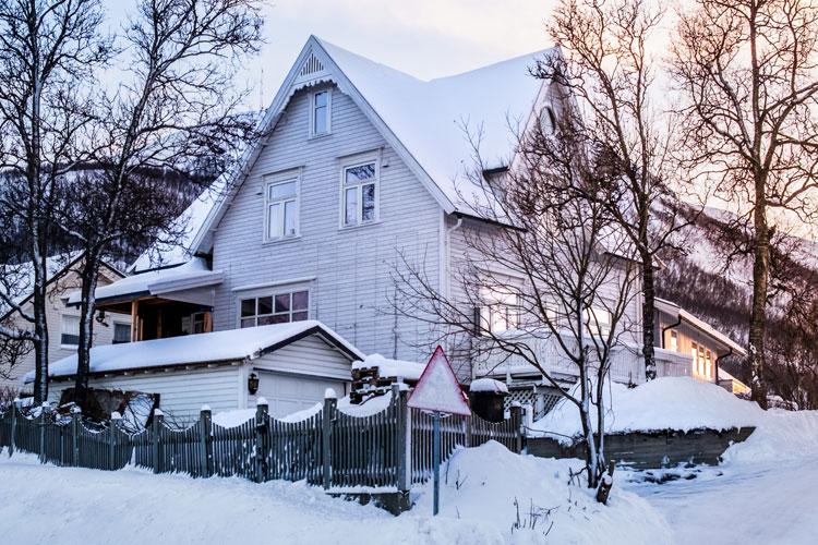 En stor, hvit trehus dekket av snø, med trær rundt og en smijernsgrind foran.