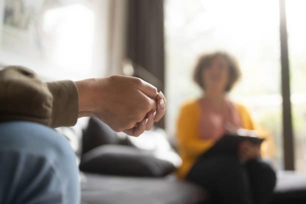 En person sitter på en sofa med en hånd som holder en ring, mens en annen person sitter i bakgrunnen med notater.