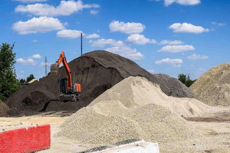 Gravemaskin som arbeider med store hauger av grus og sand under en blå himmel med skyer.