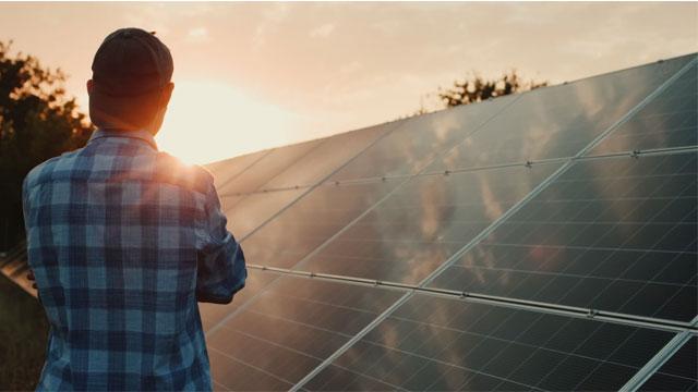 En person i rutete skjorte står foran solcellepaneler mens solen går ned i bakgrunnen.