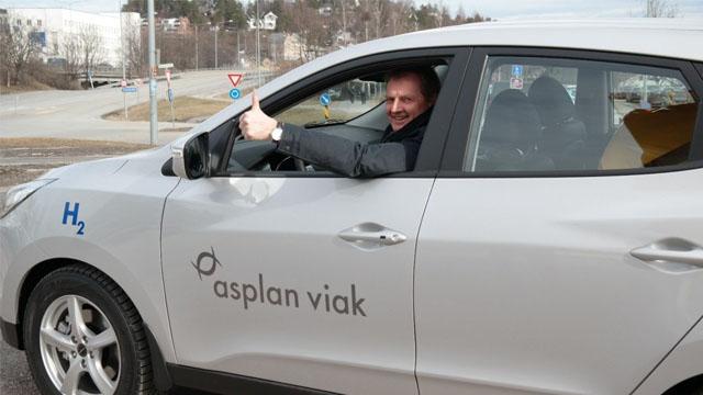 En mann smiler og gir tommel opp fra førersetet i en sølvfarget bil med "asplan viak" logo.