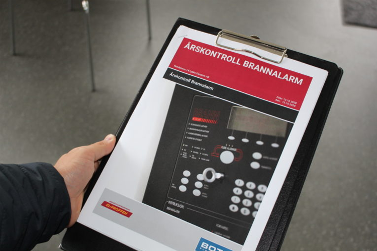 Hånd som holder en klippemappe med dokumentasjon om brannalarmkontroll. Skjermbilde av brannalarmsystemet er synlig.