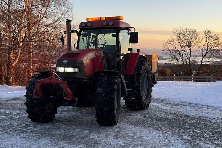 Rød traktor med snøplog står på en isete vei med trær og vann i bakgrunnen under solnedgang.