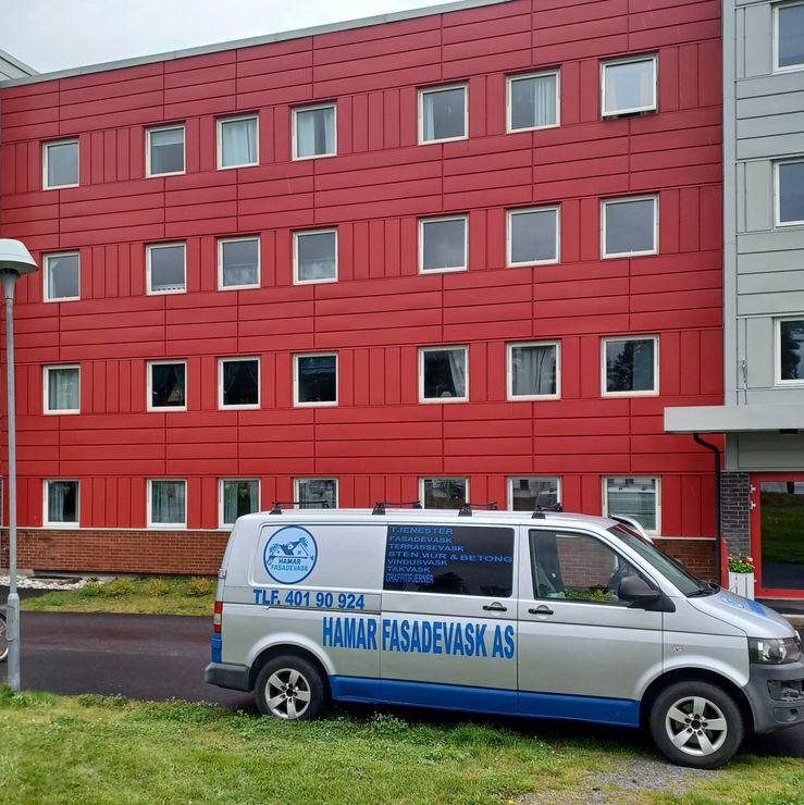 En hvit varebil med blå tekst står foran en rød og grå bygning med vinduer.