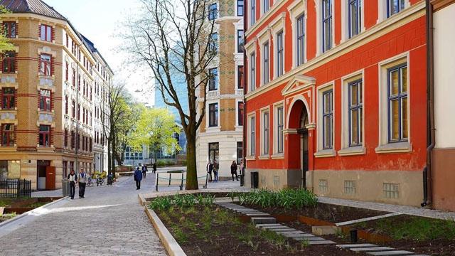 En gate med moderne bygninger og en rød bygning, omgitt av trær og folk som går forbi.