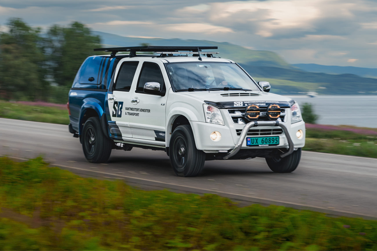 En modifisert SUV kjører raskt på en vei med grønt landskap og fjell i bakgrunnen.