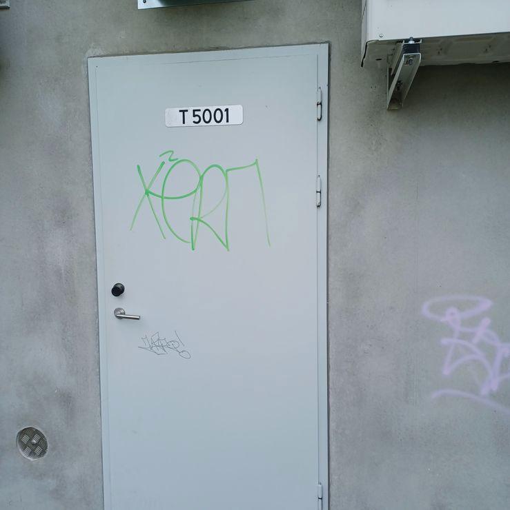 En grå dør med graffiti og nummeret T5001 på en vegg.