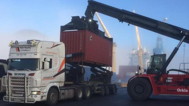 Lastebil med container heves av kran på havnen, med industrielle bygninger i bakgrunnen.