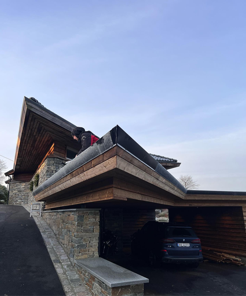 Moderne hus med skråtak og steinvegg, en person på taket og en bil parkert under.