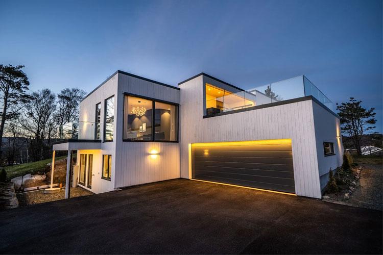 Moderne enebolig med stor terrasse og garasje, belyst av varmt lys ved skumring.