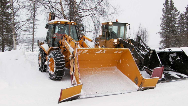 To store traktorer med snøploger i vinterlandskap, dekket av snø og trær i bakgrunnen.