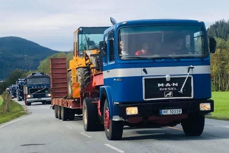 En blå MAN lastebil med en traktor på tilhenger, kjører på en vei med grønne åser i bakgrunnen.