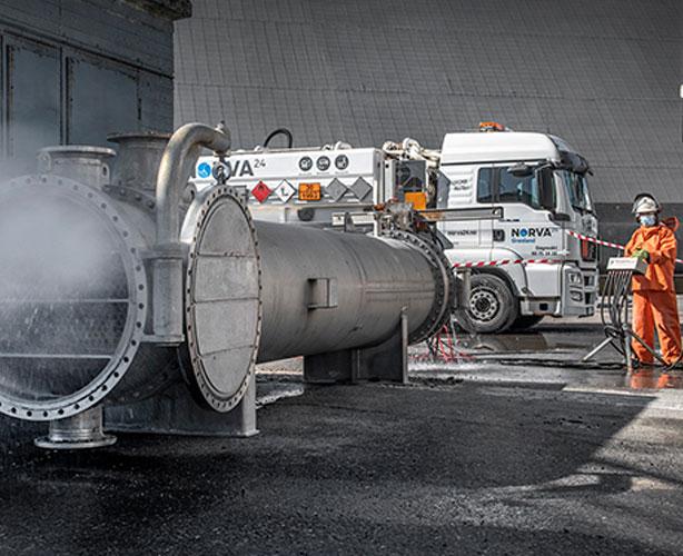 En stor industrirørledning med damp som kommer ut, med en lastebil i bakgrunnen og en arbeider i oransje drakt.