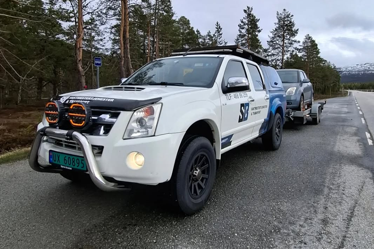 En hvit SUV med takstativ og lysbøyle, som trekker en blå bil på tilhenger, parkert langs en vei i naturen.