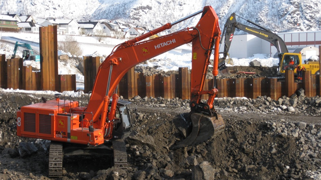 Gravemaskin i arbeid på en byggeplass med snø og fjell i bakgrunnen.