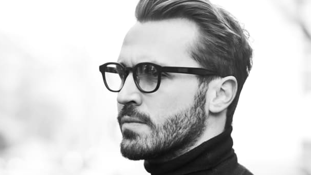 En mann med briller og skjegg, iført en svart genser, står i profil mot en lys bakgrunn.