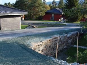 En vei med grus og steinmur, omgitt av trær og røde hytter i bakgrunnen.