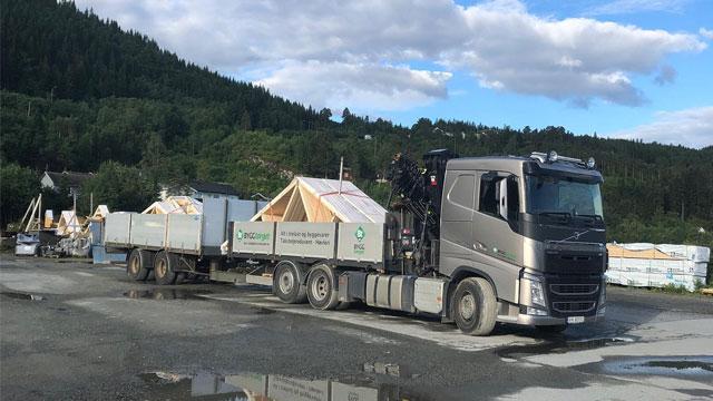 En lastebil med tilhenger som frakter byggevarer på en byggeplass omringet av trær og fjell.